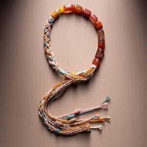 May include: A handcrafted necklace featuring a strand of reddish-brown, square-cut beads. The necklace has a braided section with pastel-colored threads and a tassel of matching threads. The necklace is displayed on a neutral background.