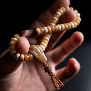 Puede incluir: Un rosario de cuentas redondas, lisas y translúcidas en tonos beige y marrón claro. El cordón incluye una cuenta cilíndrica de ágata con bandas marrones y blancas, una pieza central tallada con textura y una borla de hilo marrón.
