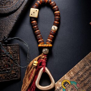 May include: A handcrafted prayer bead necklace with a wooden bead design. The necklace features ivory-colored square beads with dot patterns, a tassel, and a triangular pendant. The beads are strung together with a red and brown cord.