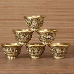 May include: A set of six small brass bowls arranged in a pyramid stack. Each bowl features a metallic gold finish and intricate embossed relief carvings of various figures and patterns around the exterior, with a flared rim and a pedestal base.