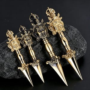 May include: Four gold-coloured metal phurba daggers arranged in a row, each featuring a detailed three-faced deity head at the handle, a cylindrical grip, and a sharp, pointed triangular blade with ornate decorative elements at the base of the hilt.