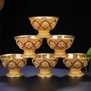 May include: A set of six gold-colored metal bowls arranged in a pyramid stack. Each bowl features a pedestal base and an exterior surface decorated with embossed lotus petal patterns highlighted by deep red enamel accents along the edges.