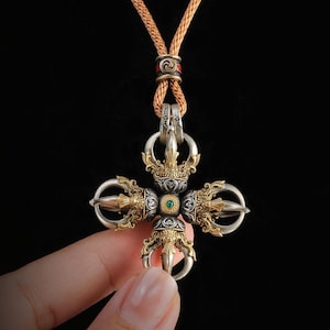 May include: A double vajra pendant featuring intricate gold and silver metalwork with a central green stone. The pendant is suspended from a braided tan cord that includes a small cylindrical bead with red and silver accents at the top.