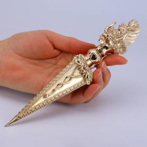 Könnte beinhalten: Ein metallisch goldfarbener Ritualdolch mit einem verzierten Griff, der von einer detaillierten Drachenkopfskulptur gekrönt wird. Die Klinge ist mit einem erhabenen, gewundenen Schlangenmotiv und komplizierten, texturierten Rändern verziert.