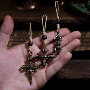 Può includere: Tre ciondoli in metallo color bronzo con intricati disegni vajra e phurba. Ogni ciondolo è fissato a un cordino intrecciato color marrone chiaro con una piccola perla marrone scuro. I componenti in metallo presentano incisioni dettagliate e finitura.