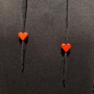 May include: Close-up of a black hoodie with red heart-shaped drawstring toggles. The hoodie's fabric has a textured appearance. The drawstring cords are black and the heart toggles are a bright red color.