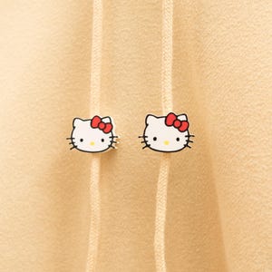 May include: Two white Hello Kitty face pins with red bows and black whiskers are attached to a beige hoodie. The pins are positioned on either side of the hoodie's drawstring, adding a touch of playful charm to the garment.