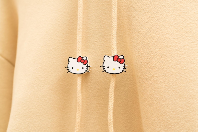 May include: Two white Hello Kitty face pins with red bows and black whiskers are attached to a beige hoodie. The pins are positioned on either side of the hoodie's drawstring, adding a touch of playful charm to the garment.