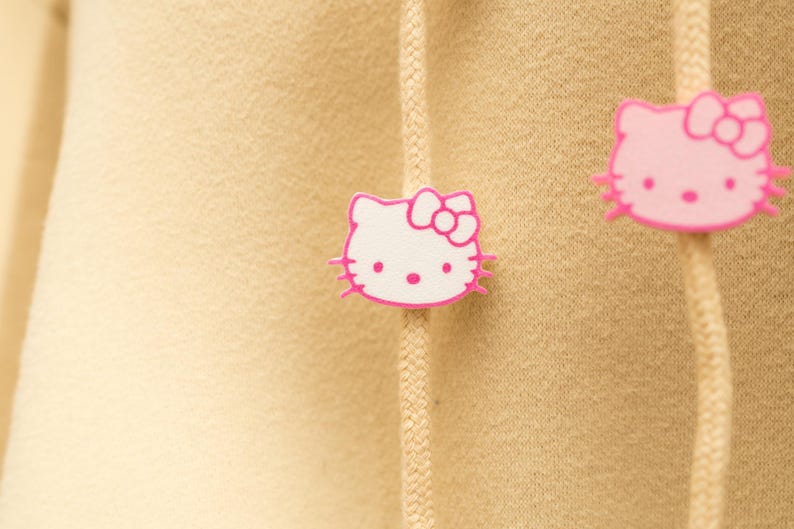 May include: Close-up of a tan hoodie with two pink and white Hello Kitty face patches. The patches are on the drawstring of the hoodie. The Hello Kitty faces have pink bows and whiskers.