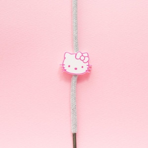 May include: A pink and white Hello Kitty charm on a gray cord against a pink background. The charm features the iconic character's face with a bow. The cord is centered vertically.