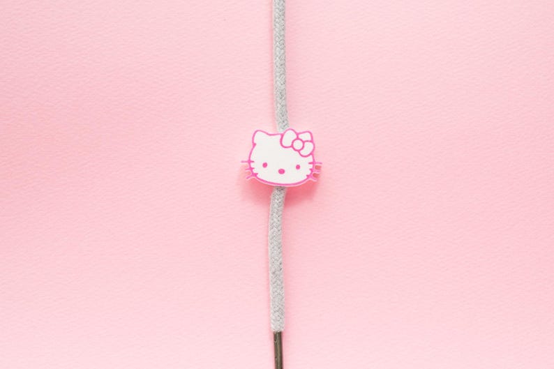 May include: A pink and white Hello Kitty charm on a gray cord against a pink background. The charm features the iconic character's face with a bow. The cord is centered vertically.