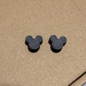 May include: Two dark gray Mickey Mouse head-shaped objects. The objects are made of a textured material and are placed on a brown surface. The objects are small and have a three-circle design.
