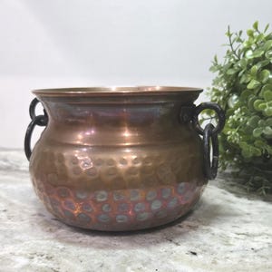 May include: A round, hammered copper pot featuring a wide, flared rim and two dark, circular metal ring handles attached to the sides. The exterior surface displays a textured, dimpled pattern across the lower half and a smooth, polished finish.