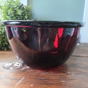 May include: A deep, translucent dark red glass mixing bowl with a smooth, rounded rim and a polished finish. The bowl features a wide, circular opening that tapers down to a smaller base, reflecting light across its curved, glossy surface.