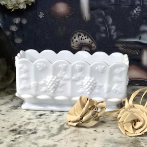 May include: A white rectangular planter with a scalloped rim and grape cluster detailing. The planter is made of opaque glass and sits on a marble surface. Decorative paper flowers are in the foreground.