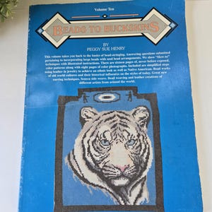 May include: A blue instructional book titled Beads to Buckskins Volume Ten by Peggy Sue Henry. The cover features a graphic illustration of a white tiger head centered within a dark rectangular frame against a bright blue background.