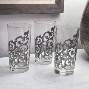 May include: Three clear drinking glasses with a decorative gray scroll design. The glasses are tall and cylindrical, with the design covering a portion of the glass. The glasses are arranged on a white surface.