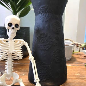 May include: A matte black cylindrical vase featuring a raised, textured floral pattern across its surface, positioned next to a small, off-white plastic skeleton figurine with black eye sockets and a neutral, smiling expression.