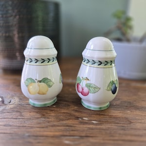 May include: Two white ceramic salt and pepper shakers with fruit illustrations. Each shaker features a green band at the base and top, with a decorative leaf pattern. One shaker has yellow fruit, the other has cherries and a plum.