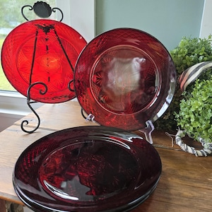 May include: A set of three round, deep red glass plates featuring a pressed diamond pattern along the outer rim and a central starburst design. One plate is displayed upright on a black metal stand, while two others are stacked horizontally.