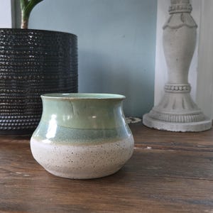 May include: A handmade ceramic vase with a speckled, off-white base, a band of muted teal, and a sage green top. The vase has a rounded shape and sits on a wooden surface. A black textured pot and a decorative pillar are in the background.