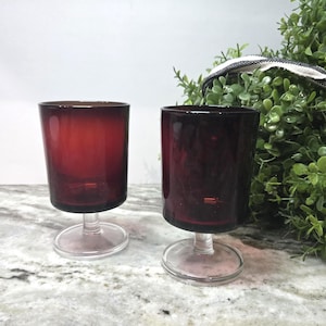 May include: Two matching stemmed glasses featuring deep burgundy red bowls and clear glass bases. The glasses have a cylindrical shape with a smooth finish and a short, sturdy stem connecting the bowl to the circular foot, creating a balanced look.