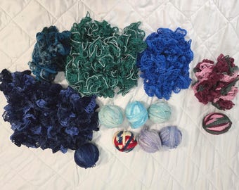 May include: A collection of various yarn types including several bundles of ruffled, decorative ribbon yarn in shades of dark blue, forest green, bright blue, and variegated pink, alongside multiple round balls of solid and multi-colored yarn.