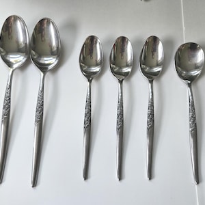 May include: A set of six silver-toned metal spoons arranged in a row. The collection includes two larger spoons and four smaller spoons, all featuring a detailed floral pattern embossed along the handle stems and polished oval bowls.
