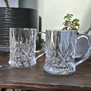 May include: Two clear crystal mugs with intricate diamond and vertical line patterns. Each mug has a curved handle. The mugs are on a wooden surface, with a large black textured pot and plants in the background.