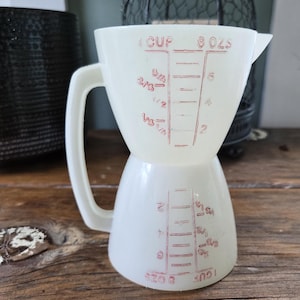 May include: A white milk glass measuring cup with a handle, featuring red printed markings for 1 cup and 8 ounces. The design includes a dual-sided scale with fractional and ounce increments, allowing for measurements when the cup is upright or inverted.