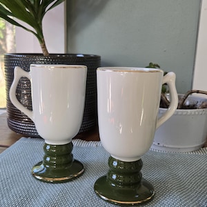 May include: Two white ceramic mugs with a dark green base and gold trim. The mugs have a unique handle design and are displayed on a blue patterned surface. A plant in a black pot is in the background.