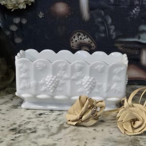 May include: A white rectangular planter with a scalloped rim and grape cluster designs. The planter is made of opaque glass and sits on a marble surface. Decorative paper flowers are in front of the planter.