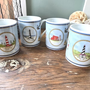 May include: Four white ceramic mugs featuring light blue backgrounds and nautical motifs. Each mug displays a central lighthouse illustration framed by a gold rope border, accented with small anchors, bells, and life preservers around the exterior.