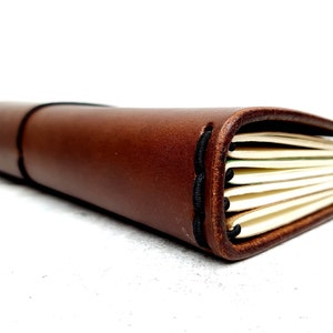 May include: A brown leather-bound journal with a stitched spine and elastic closure. The journal is closed and lying on a white surface.
