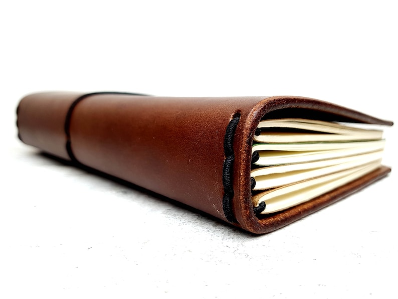 May include: A brown leather-bound journal with a stitched spine and elastic closure. The journal is closed and lying on a white surface.