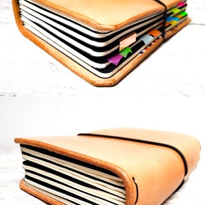 May include: A tan leather journal with black and white lined pages. The journal is secured with a black elastic band and has colorful page markers. A pen rests on top. The journal is closed in the second image.