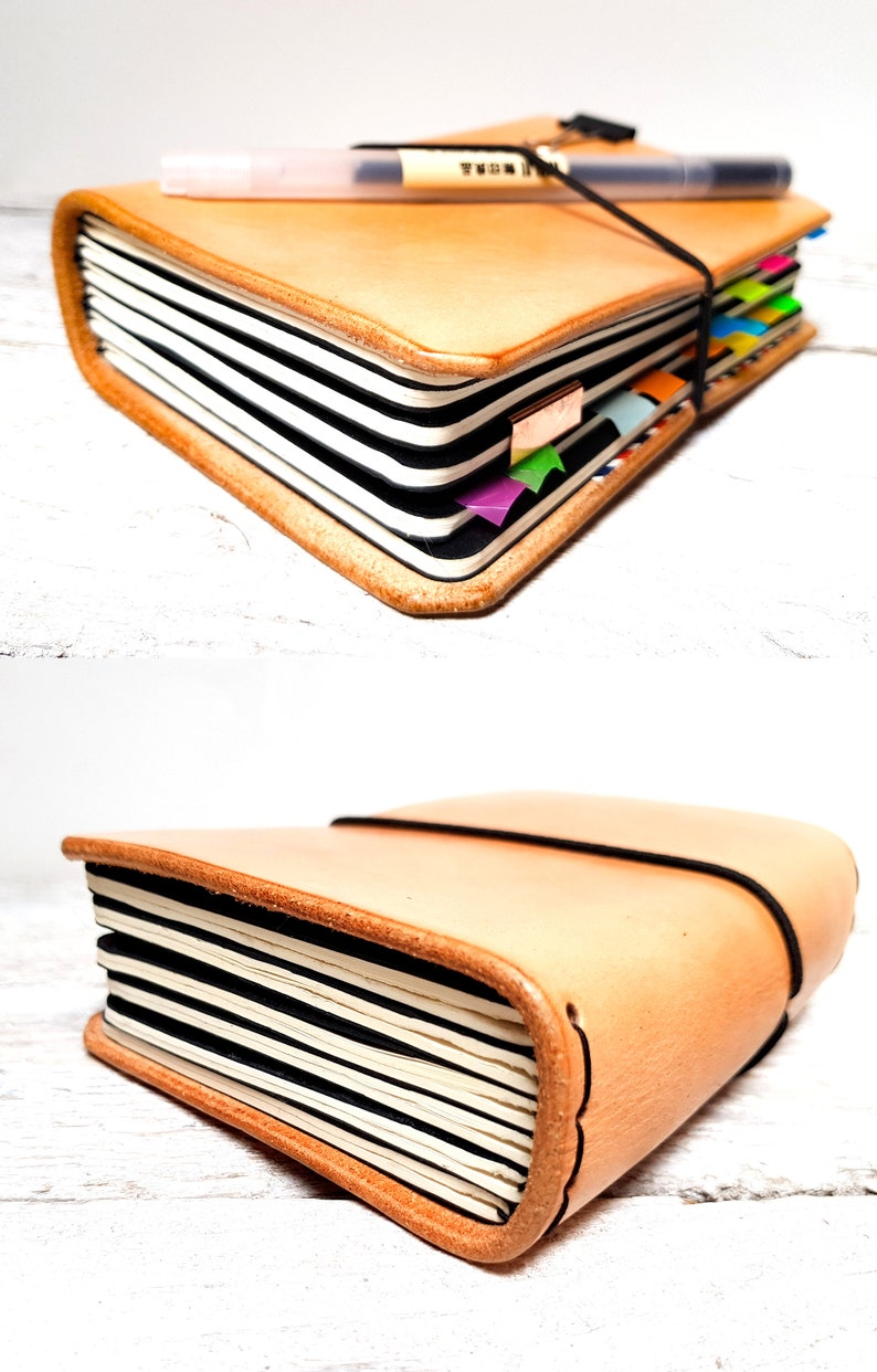 May include: A tan leather journal with black and white lined pages. The journal is secured with a black elastic band and has colorful page markers. A pen rests on top. The journal is closed in the second image.