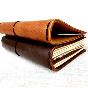 May include: Two leather-bound journals, one brown and one dark brown, stacked on top of each other. The top journal is open, revealing blank pages.
