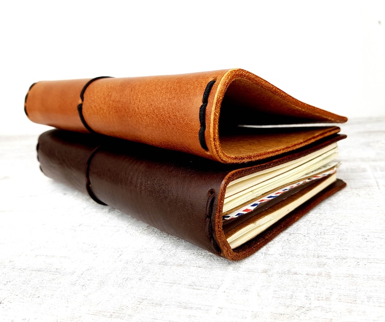 May include: Two leather-bound journals, one brown and one dark brown, stacked on top of each other. The top journal is open, revealing blank pages.