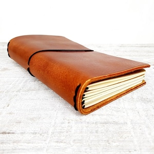 May include: A brown leather journal with a black elastic closure. The journal has a stitched spine and a flap closure. The pages are cream-colored and visible. The journal is rectangular and appears to be handmade.