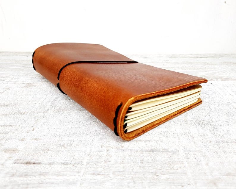 May include: A brown leather journal with a black elastic closure. The journal has a stitched spine and a flap closure. The pages are cream-colored and visible. The journal is rectangular and appears to be handmade.
