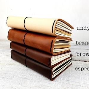 May include: Four leather-bound journals stacked on top of each other. The journals are in different shades of brown, from light to dark. The colours are labelled "undyed", "brandy", "brown", and "espresso".