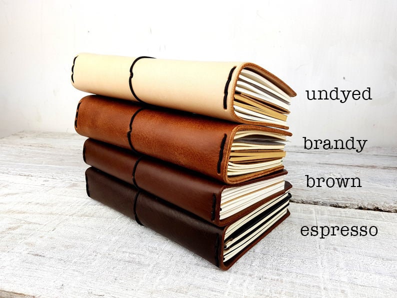 May include: Four leather-bound journals stacked on top of each other. The journals are in different shades of brown, from light to dark. The colours are labelled "undyed", "brandy", "brown", and "espresso".