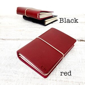 May include: A red leather journal with a cream elastic band and the initials "LH" embossed on the cover. A black journal is visible in the background, with the word "Black" written in black text. The word "red" is written in red text.