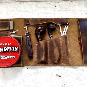 Puede incluir: Un rollo de pipa de cuero marr&oacute;n con m&uacute;ltiples bolsillos y compartimentos. El rollo est&aacute; abierto y contiene dos pipas, una lata de tabaco etiquetada "Lloyd's Bondman Rich Dark Virginia Blend", un llavero y suministros de limpieza para pipas.