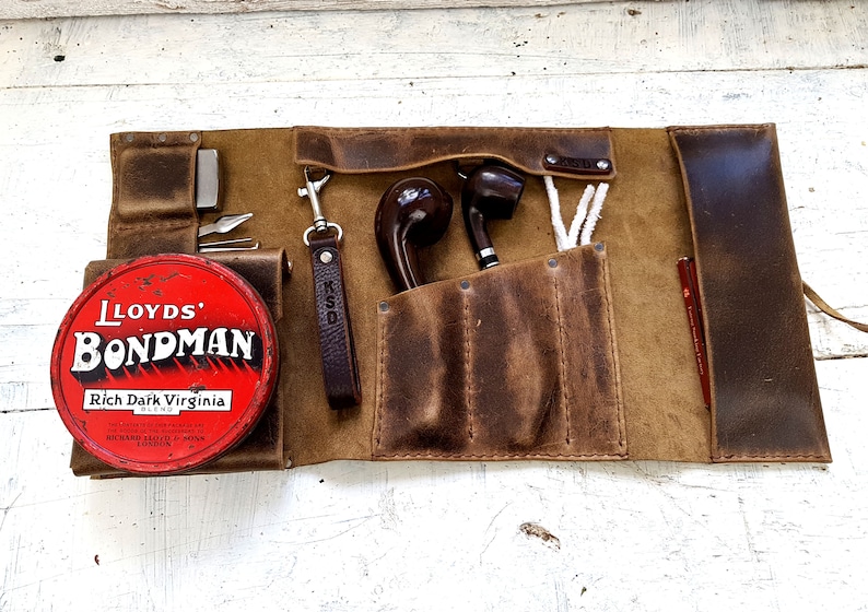 Puede incluir: Un rollo de pipa de cuero marr&oacute;n con m&uacute;ltiples bolsillos y compartimentos. El rollo est&aacute; abierto y contiene dos pipas, una lata de tabaco etiquetada "Lloyd's Bondman Rich Dark Virginia Blend", un llavero y suministros de limpieza para pipas.