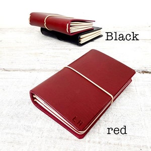 May include: A red leather journal with a cream-colored elastic band and the initials "LH" embossed on the cover. A black journal is visible in the background, with the word "Black" written in black text. The word "red" is written in red text.