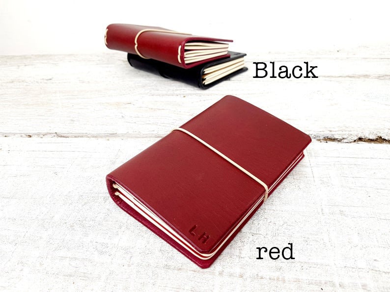 May include: A red leather journal with a cream-colored elastic band and the initials "LH" embossed on the cover. A black journal is visible in the background, with the word "Black" written in black text. The word "red" is written in red text.