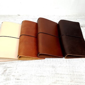 May include: Four leather journals in cream, tan, brown, and dark brown. Each journal is secured with a black elastic band. The journals are displayed side by side, showing the different colour options.