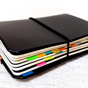 May include: A black leather notebook with a black elastic band. The pages are marked with colourful sticky tabs in orange, green, pink, and blue. The notebook is on a white wooden surface, with a light wood grain.
